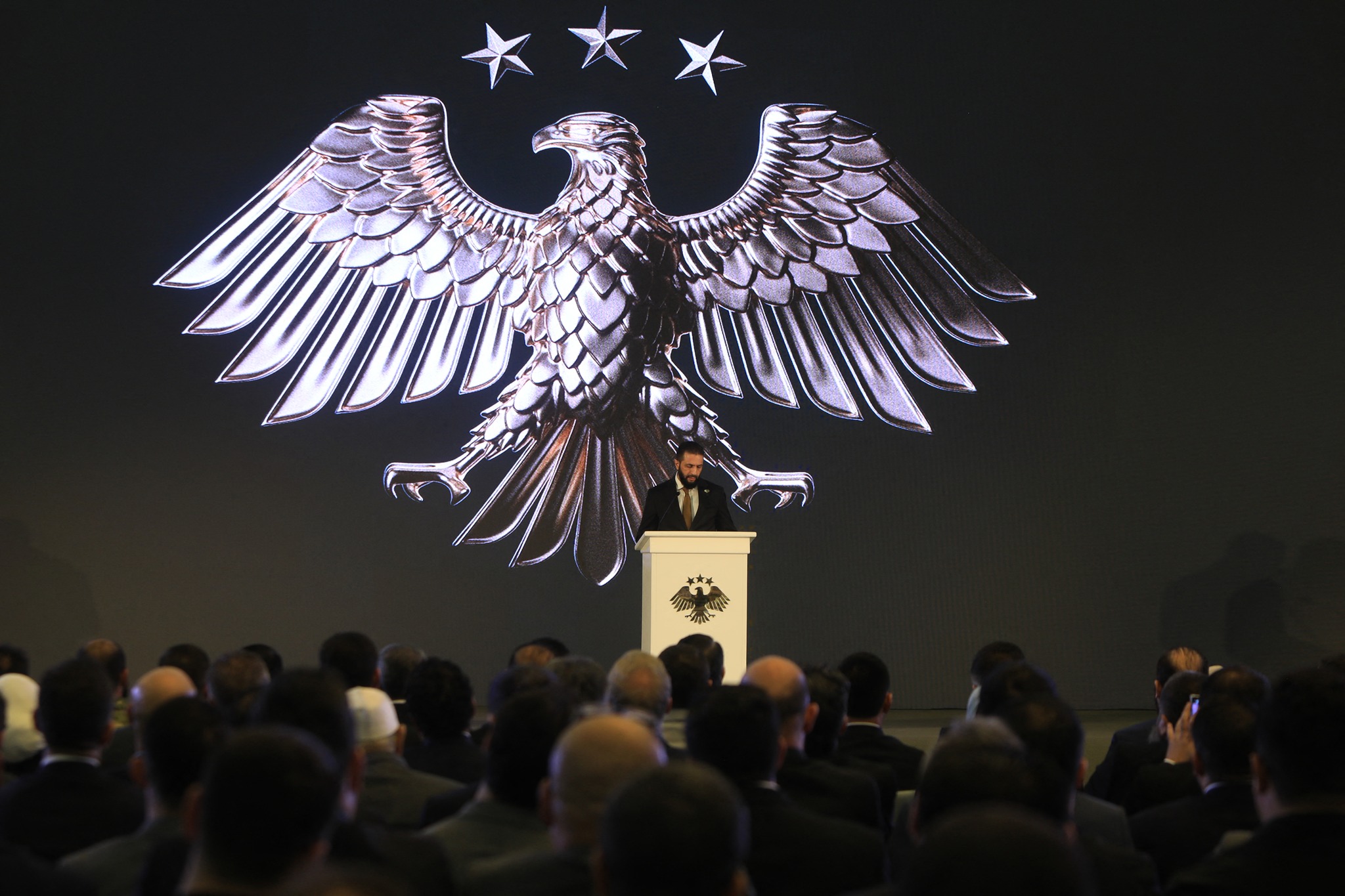 Syria's interim President Ahmad Al-Sharaa delivers a speech during the unveiling of the country's new national emblem depicting an eagle inspired by ancient motifs at Palmyra carrying 14 feathers symbolizing the country's 14 governorates and surmounted by three stars from the national flag, at the presidential palace in Damascus on July 3, 2025. (Photo by Bakr ALKASEM / AFP)