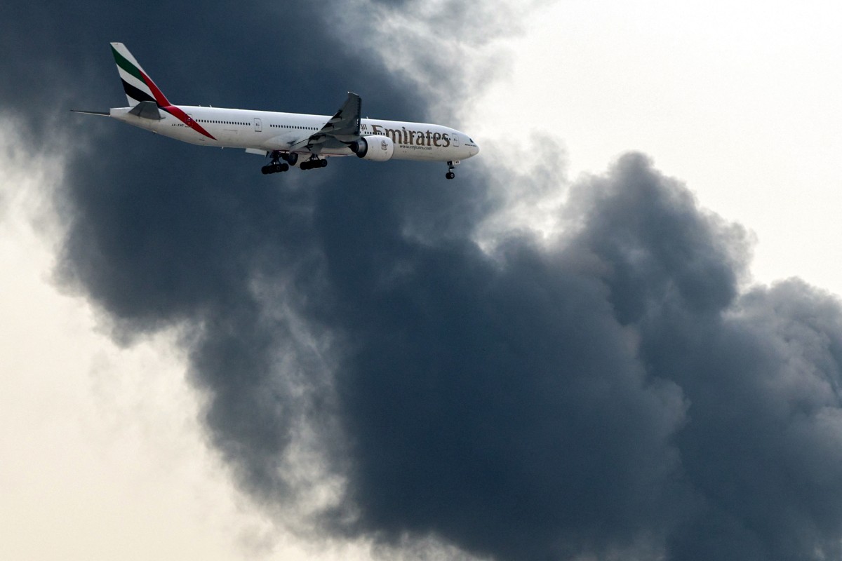 An Emirates Boeing 777 aircraft prepares for landing as a smoke plume rises from an ongoing fire near Dubai International Airport in Dubai on March 16, 2026.  (Photo by AFP)  