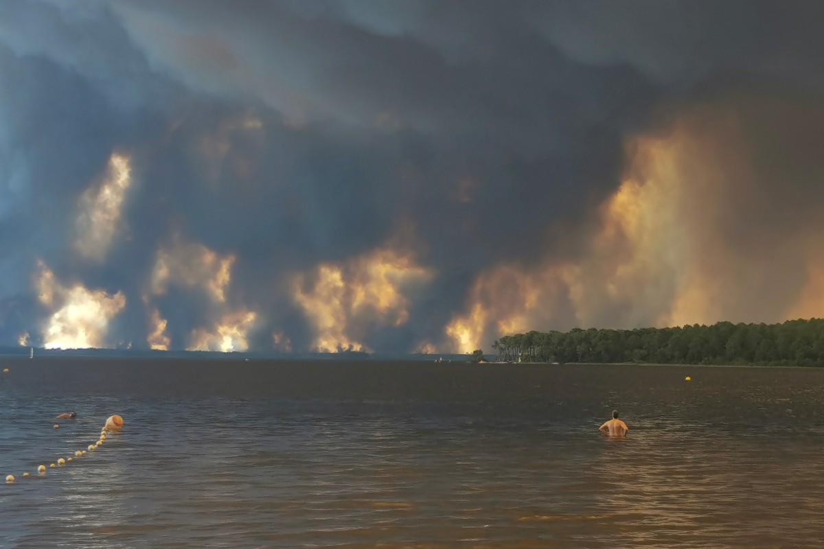 A beach goer stands in lake Lacanau, also called Etang de Lacanau (Lacanau Pond) from the beach of Moutchic in Lacanau, southwestern France on July 24, 2026, as clouds of smoke rise into the sky from a wildfire.