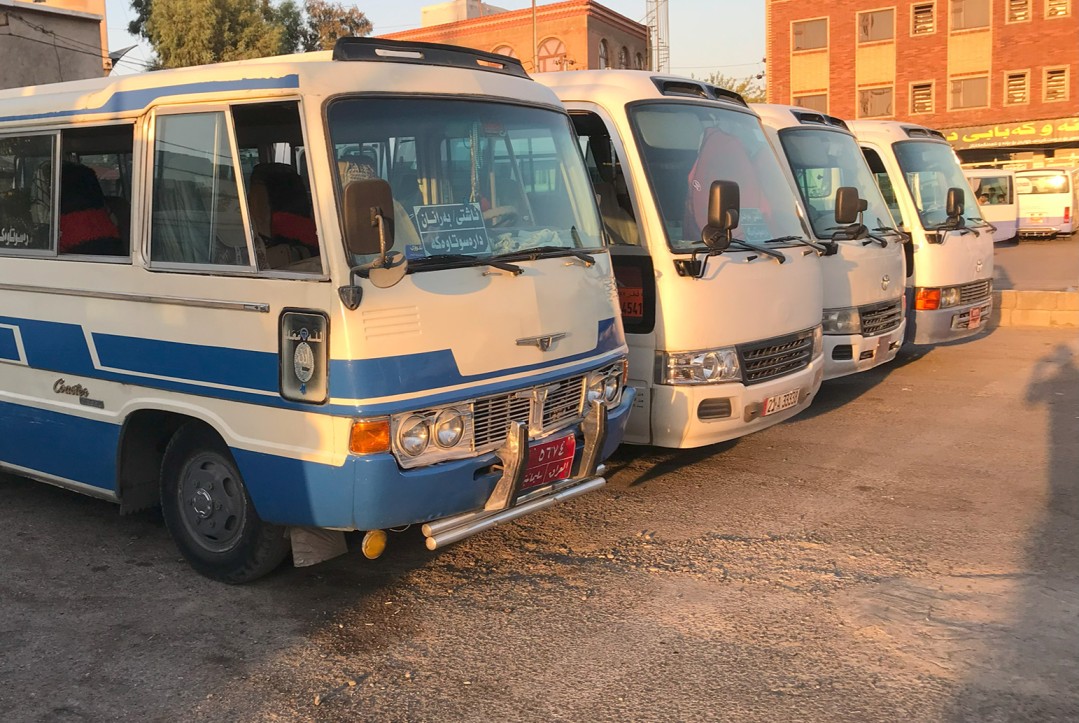 The image shows a row of four public transport mini-buses, commonly known as "coasters," parked at a transit station in Slemani.