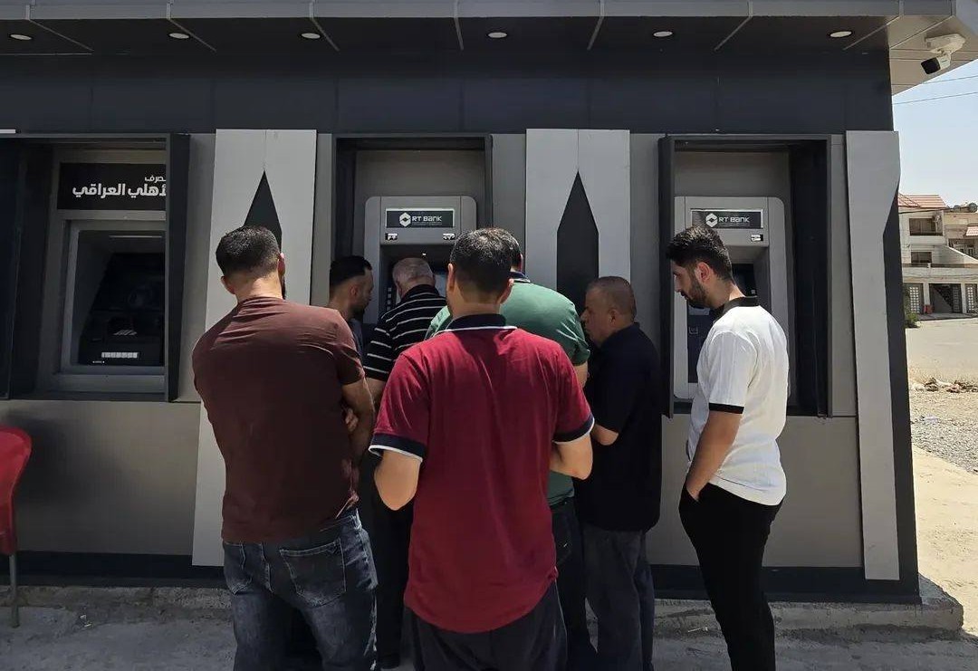 چەند هاونیشتمانییەک لە بەردەم ئامێرێکی ئەی تی ئێم دان و چاوەڕێی وەرگرتنی مووچەکانیانن