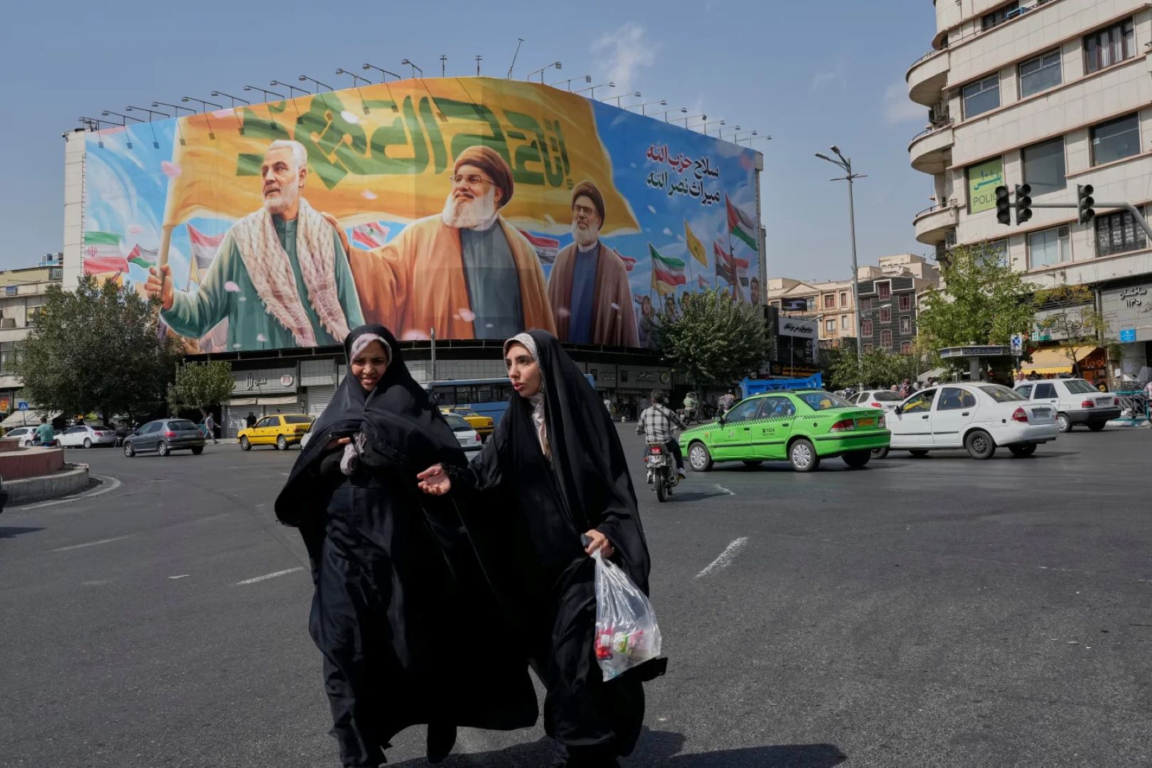 دو زن در میدان انقلاب اسلامی تهران، ایران، شنبه، ۲۷ سپتامبر ۲۰۲۵، از کنار بنر بزرگی عبور می‌کنند که تصویر قاسم سلیمانی فرماندە پیشین نیروی قدس سپاه پاسداران ایران، که در حمله پهپادی آمریکا در سال ۲۰۲۰ کشته شد و حسن نصرالله و هاشم صفی‌الدین، که در حملات هوایی اسرائیل در سال ۲۰۲۴ کشته شدند، را نشان می‌دهد. (عکس از آسوشیتدپرس/وحید سالمی)